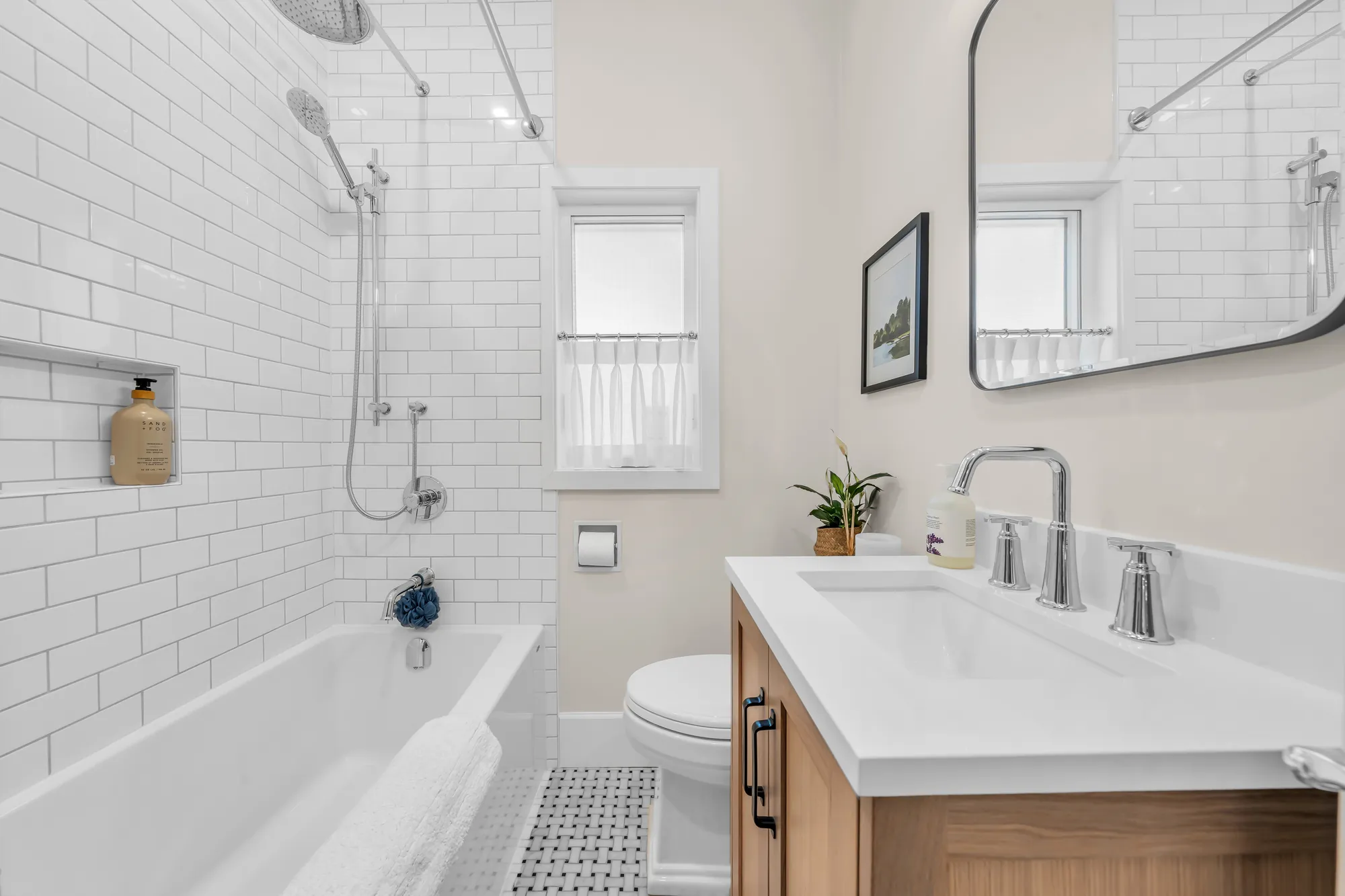 The bathroom: a tub with a white subway-tile surround and a hand shower, a wood vanity with a white top, a black-framed mirror, a mosaic floor