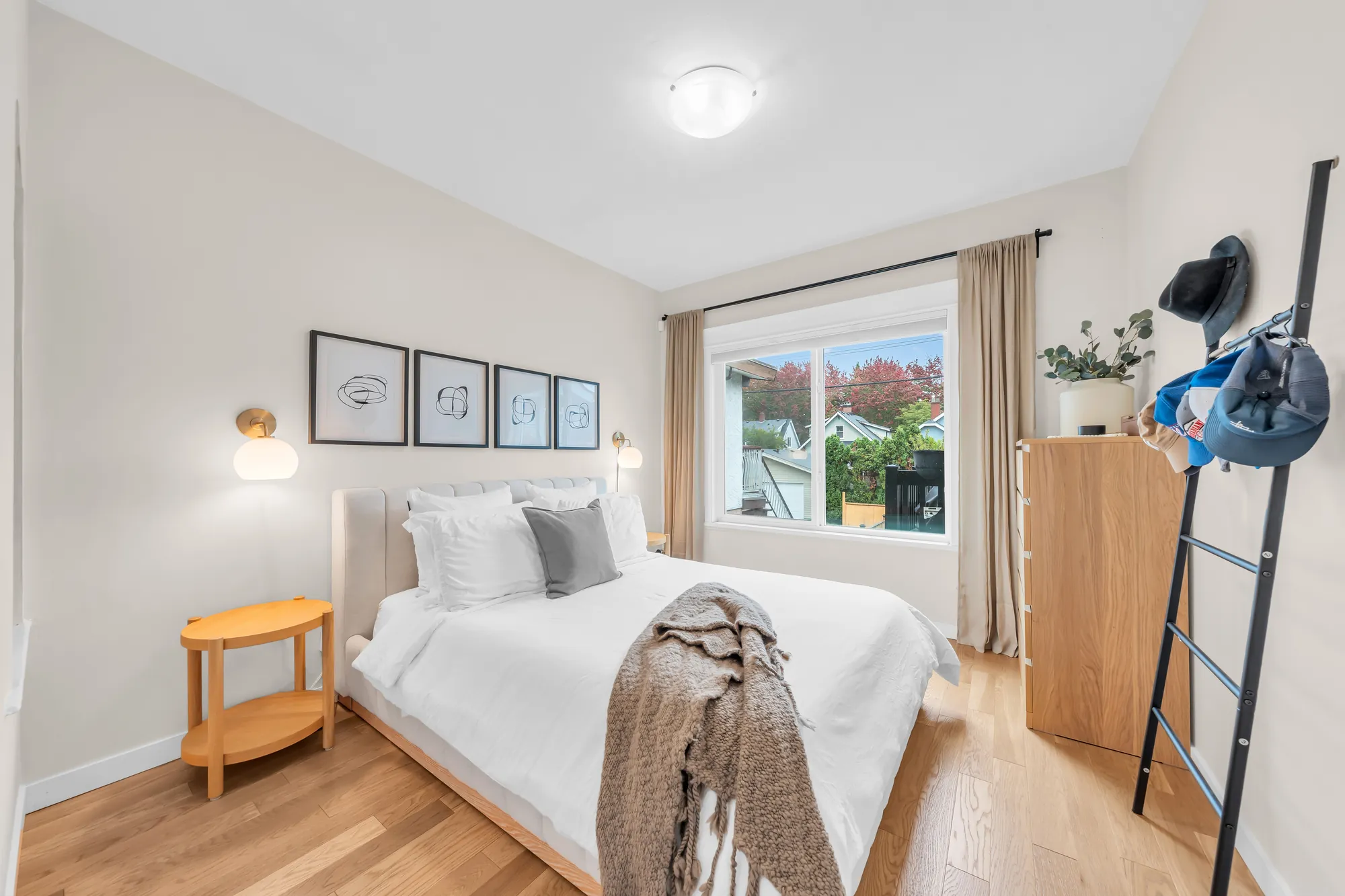The front bedroom: a bed with white linen, four framed prints, a wood side table, a ladder shelf, a window with curtains