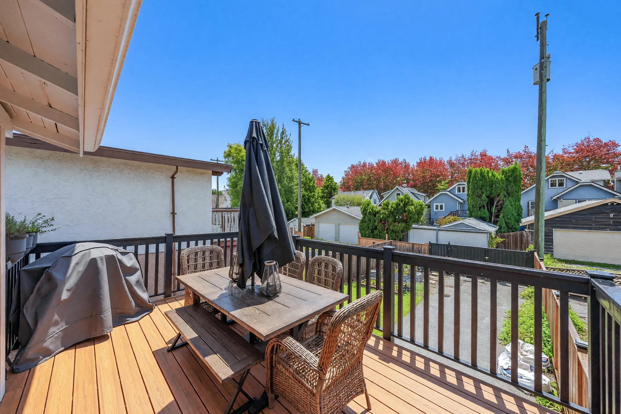 The deck: a wood table with a bench and wicker chairs, a closed umbrella, a covered barbecue, the lane and the garages beyond the railing