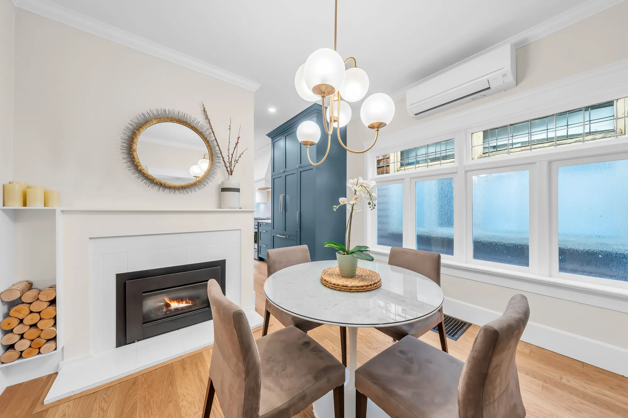 The dining room: a gas fireplace in a white mantel, a round white table with four chairs under a globe chandelier, windows along the side wall