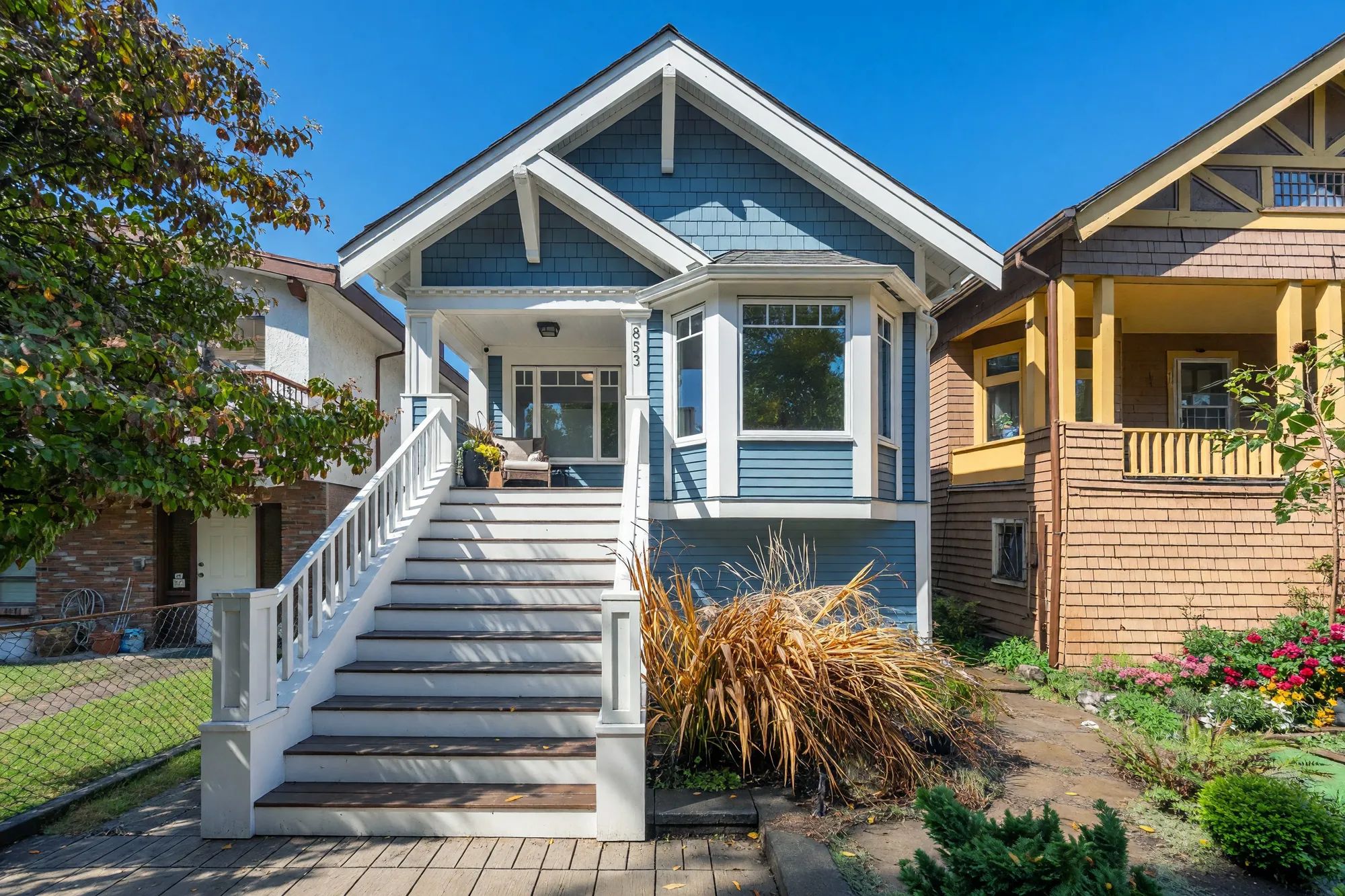 The front of the house in sunlight: blue shingle and lap siding with white trim under a steep gable, a tall white stair up to the covered porch, a bay window at the right, ornamental grasses in the front garden