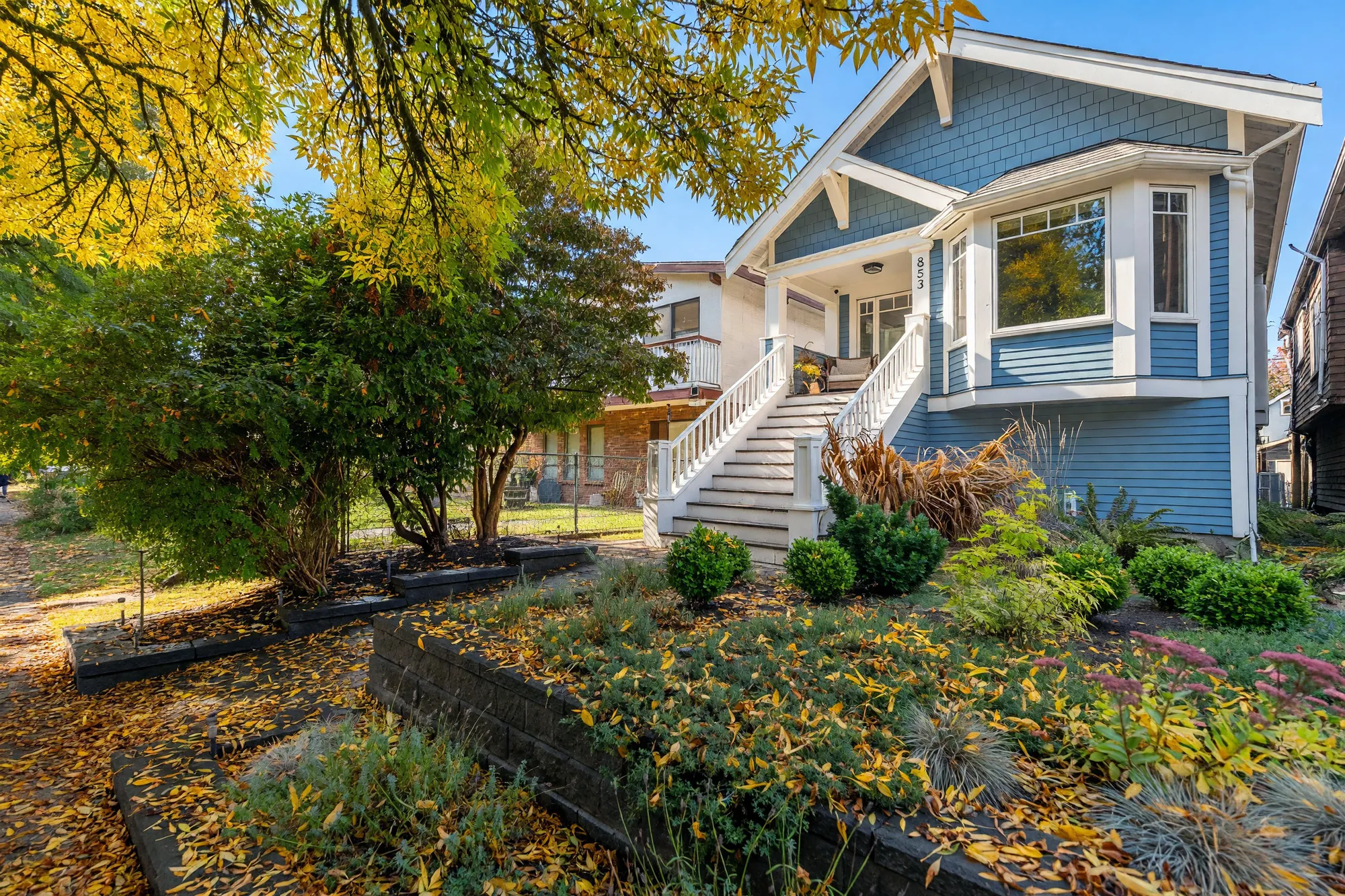 The front from the corner of the garden under a tree: the bay window and the white stair, shrubs and grasses in a bed behind a low retaining wall, fallen leaves