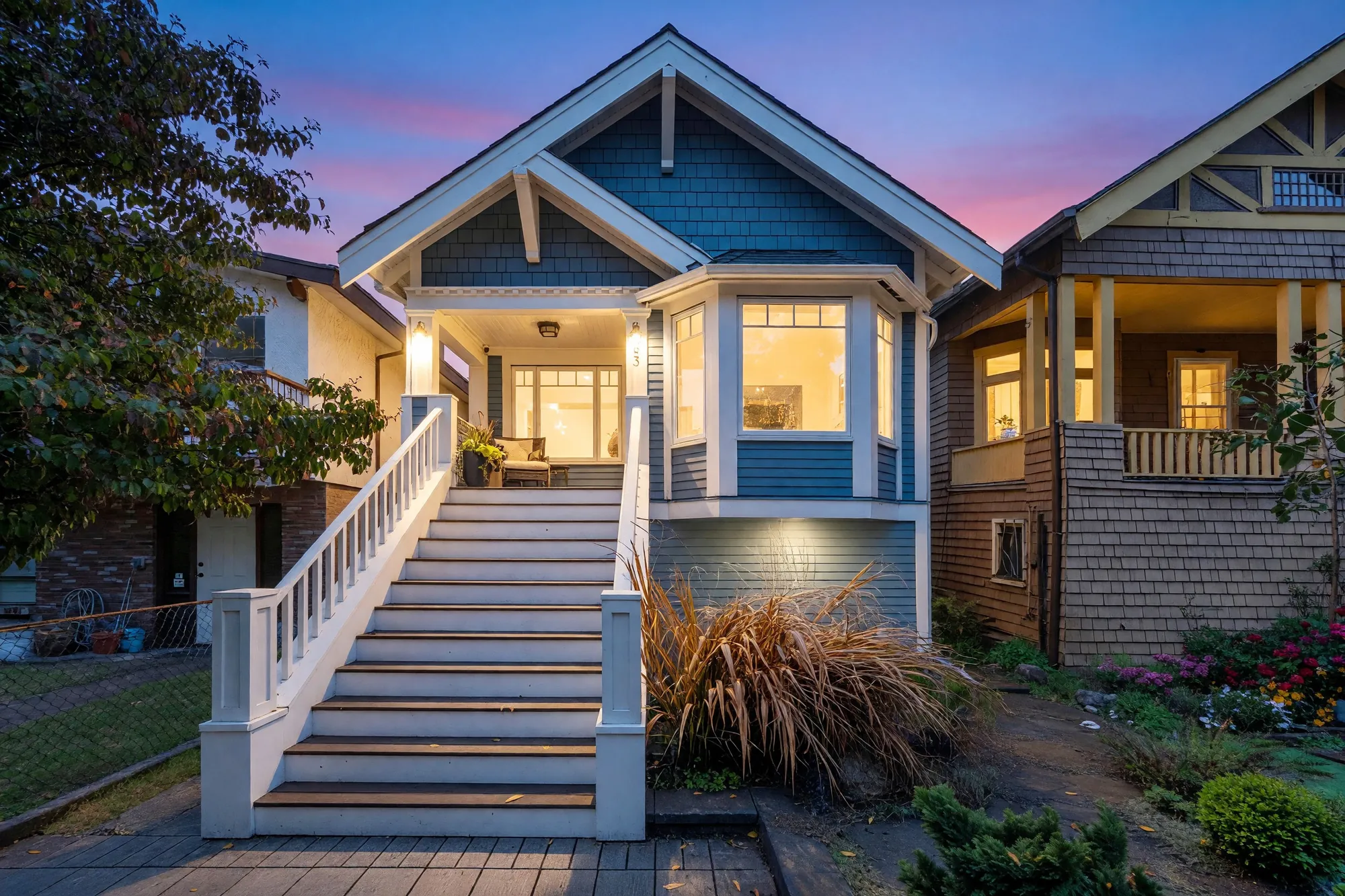The front of the house at dusk: blue siding with white trim under a steep gable, the porch light and the windows lit, a tall white stair up to the porch, a pink and violet sky