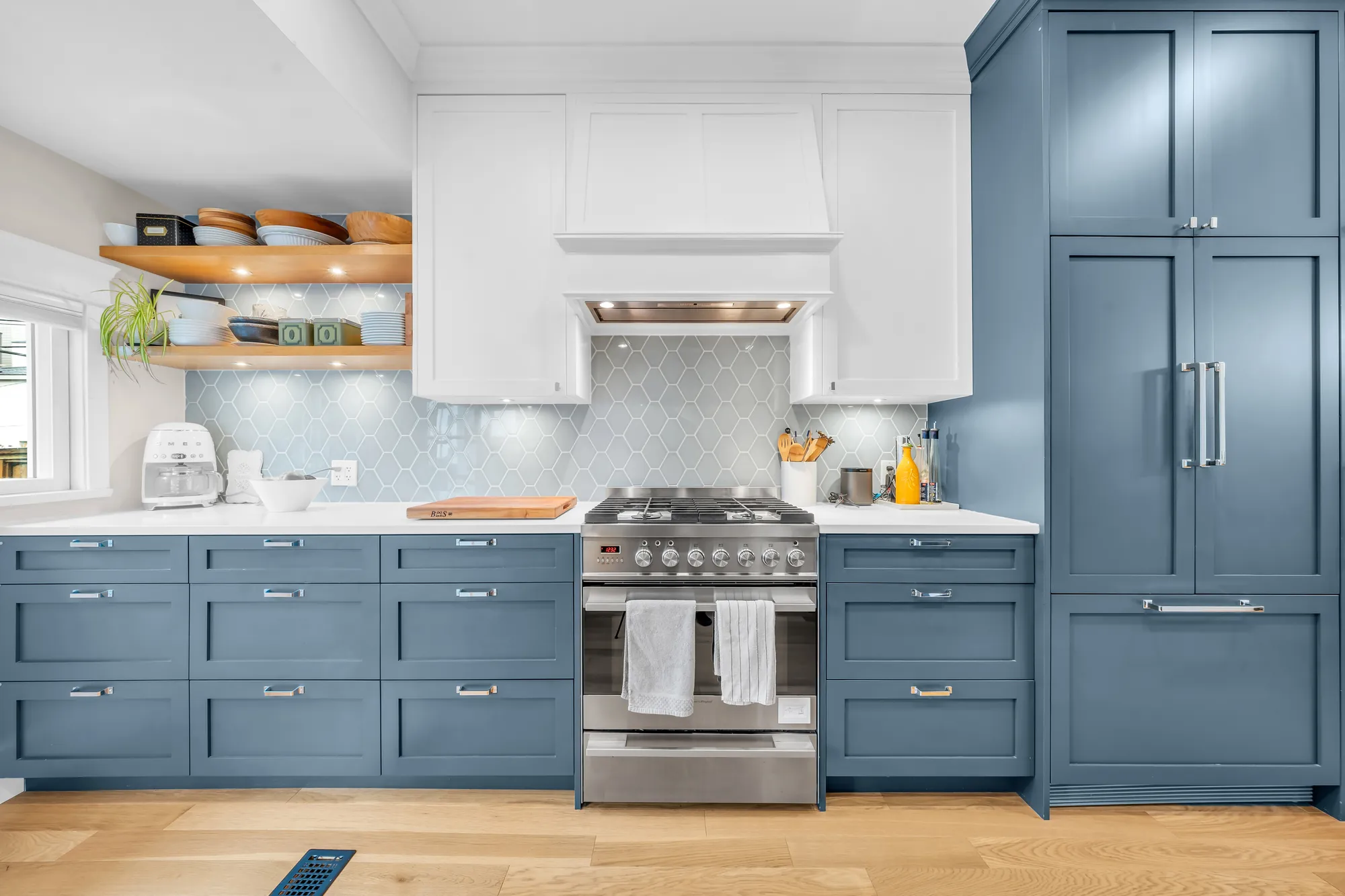 The kitchen: blue shaker cabinets, a white range hood over a stainless range, open wood shelves, blue-grey hexagon tile, a built-in fridge panelled in blue