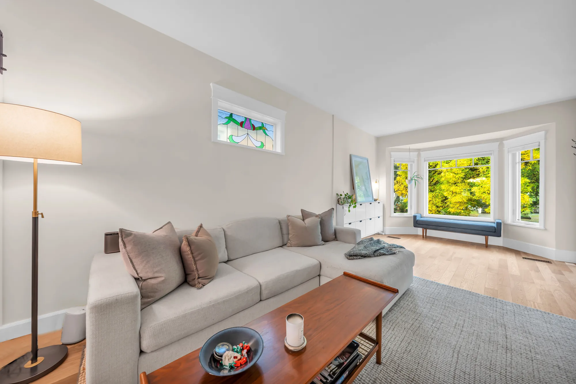 The living room toward the bay window: a grey sectional sofa, a wood coffee table, a stained-glass window high on the wall, a bench in the bay, oak floors