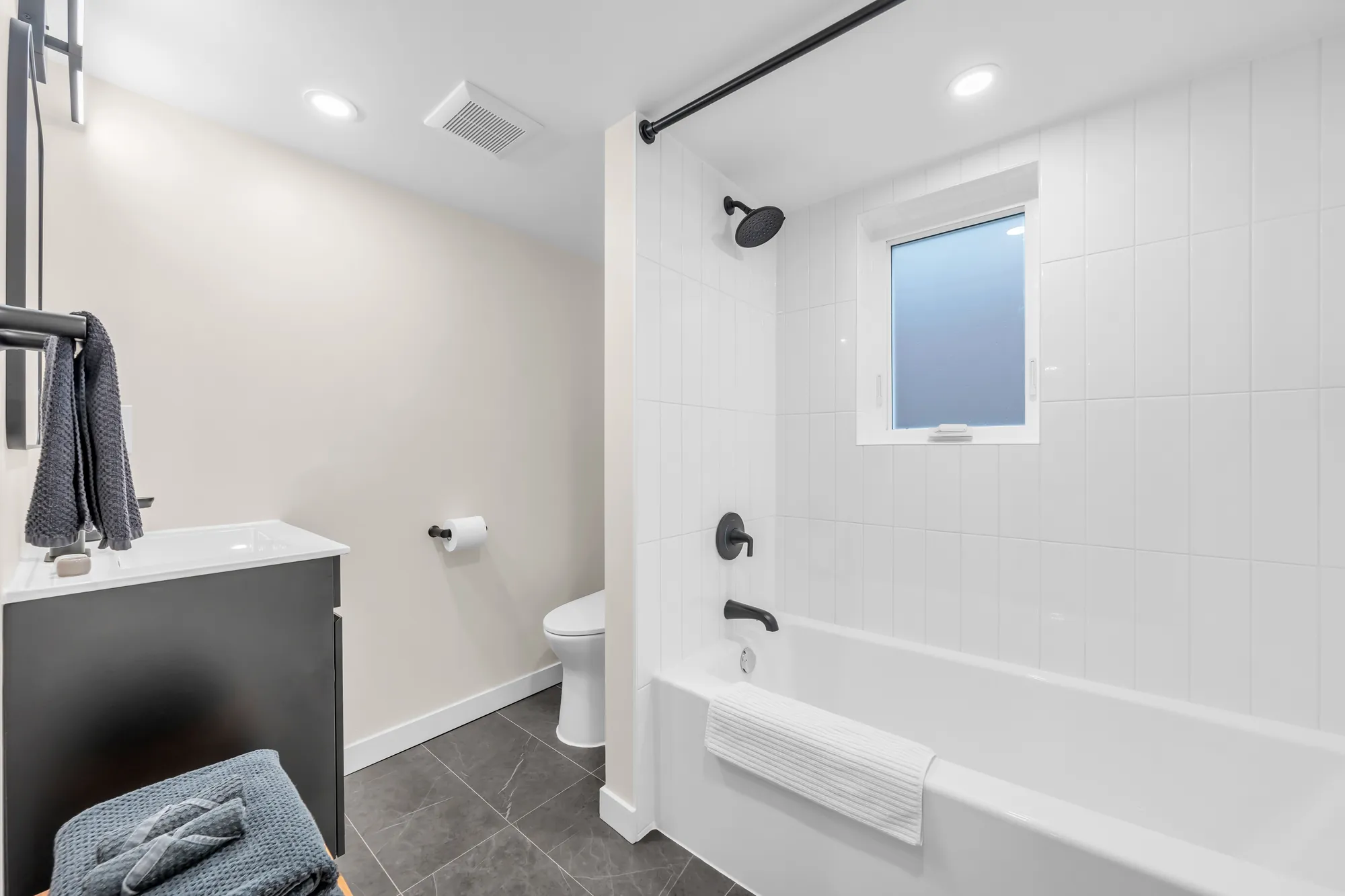 The lower bathroom: a tub with a white tile surround, a dark vanity, a toilet, a window
