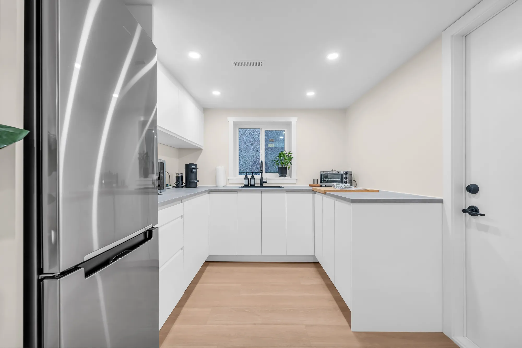 The lower kitchen: white flat-front cabinets, a stainless fridge, an L-shaped counter under a window
