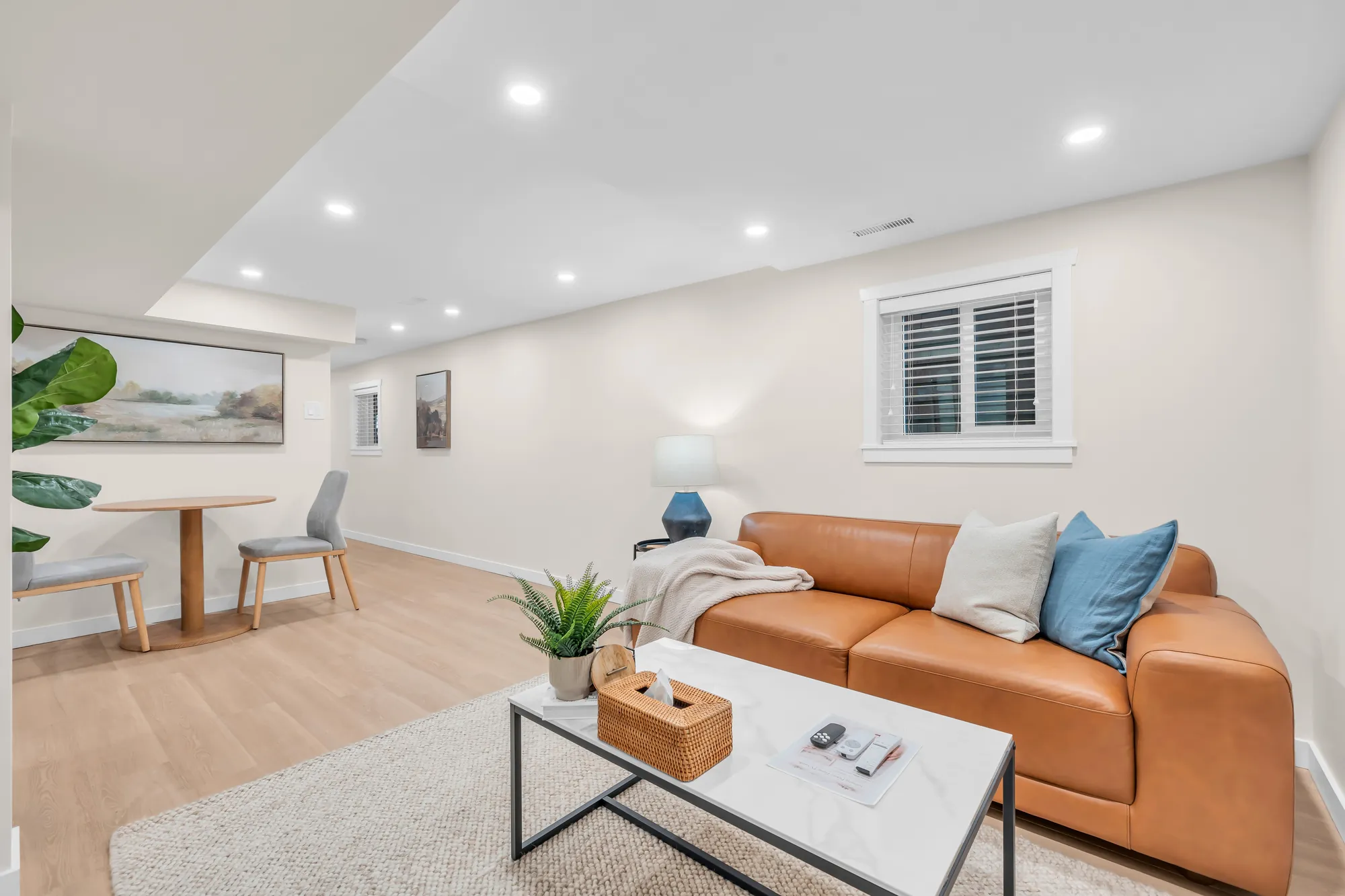 The lower floor&rsquo;s living room: a tan leather sofa, a coffee table, a round dining table at the left, a shuttered window