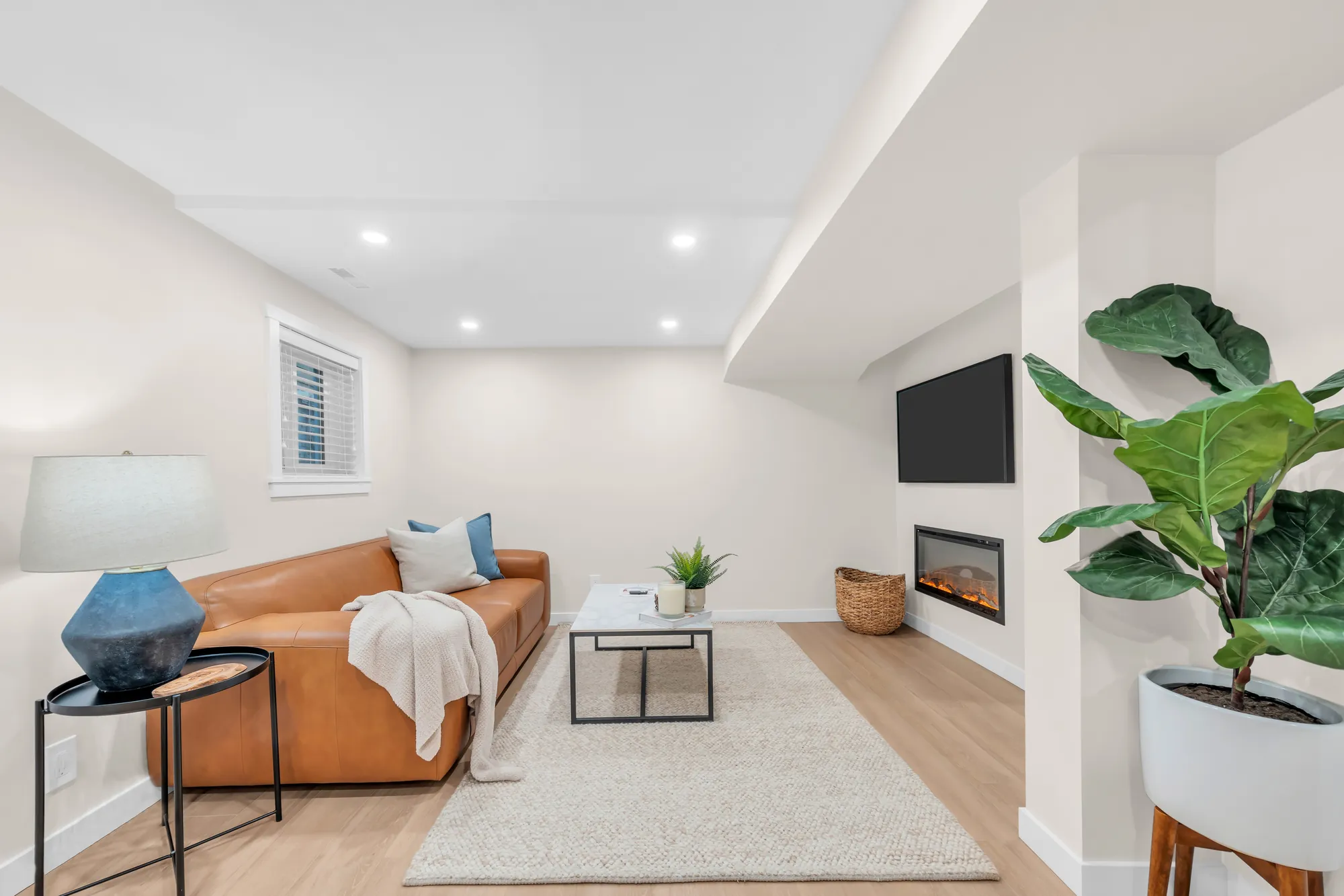 The lower living room: an electric fireplace under a wall-mounted television, a fiddle-leaf fig, the sofa and a lamp