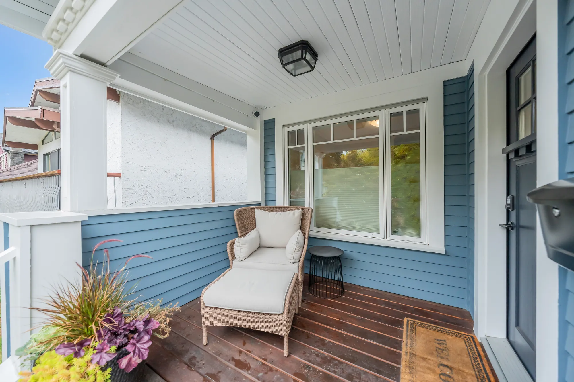 The covered porch: a wicker lounge chair and footstool on a wood floor, the front door and a wide window in blue siding, a planter of grasses