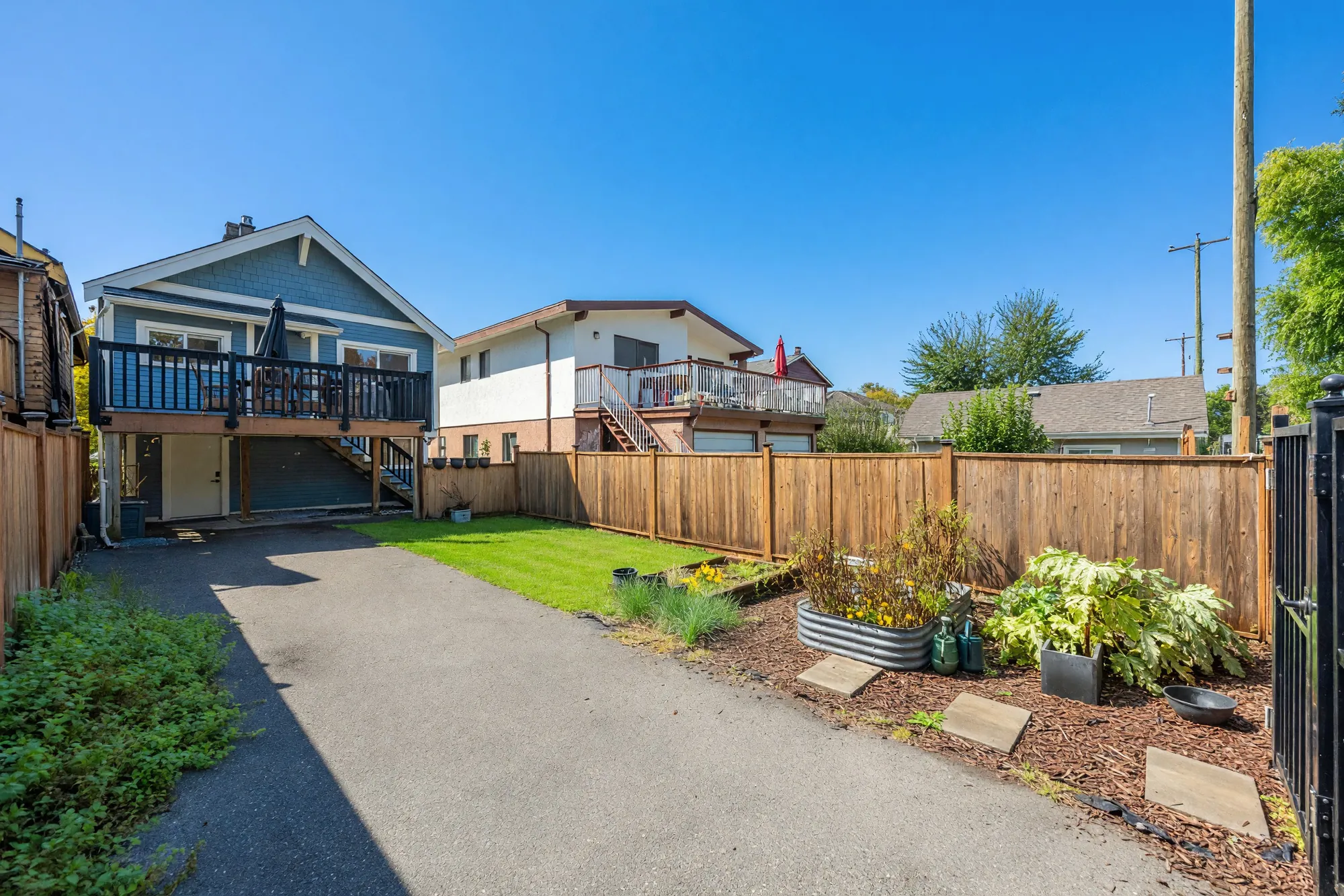 The back yard toward the house: a paved parking pad along the fence, a lawn, raised planting beds, the deck above the lower floor